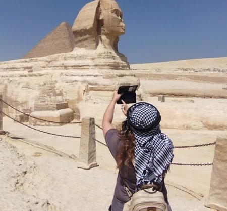 Une femme de dos portant un keffieh traditionnel photographie le Grand Sphinx de Gizeh avec son smartphone, une étape clé du voyage Pyramides depuis Sharm El-Sheikh