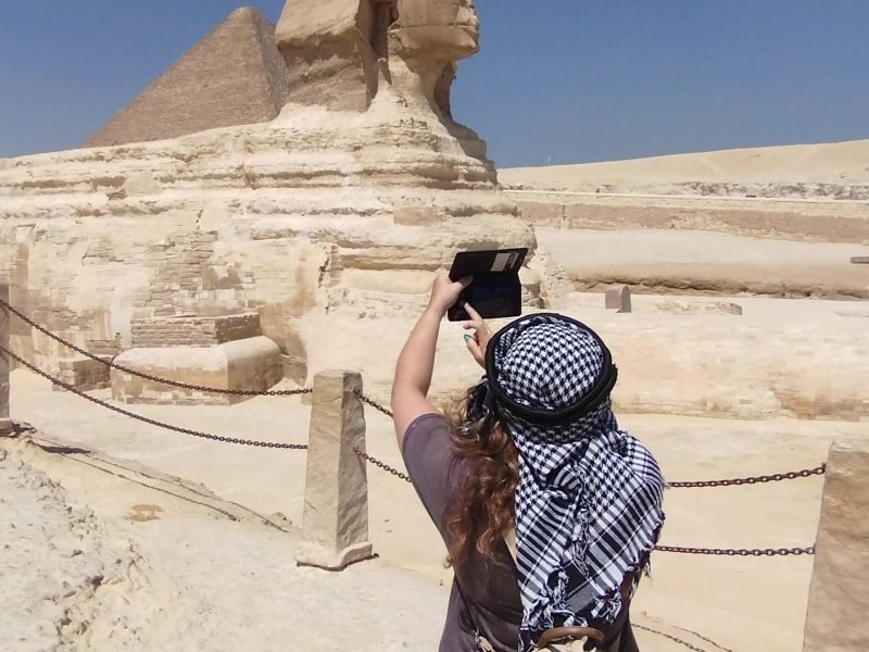 Une femme de dos portant un keffieh traditionnel photographie le Grand Sphinx de Gizeh avec son smartphone, une étape clé du voyage Pyramides depuis Sharm El-Sheikh