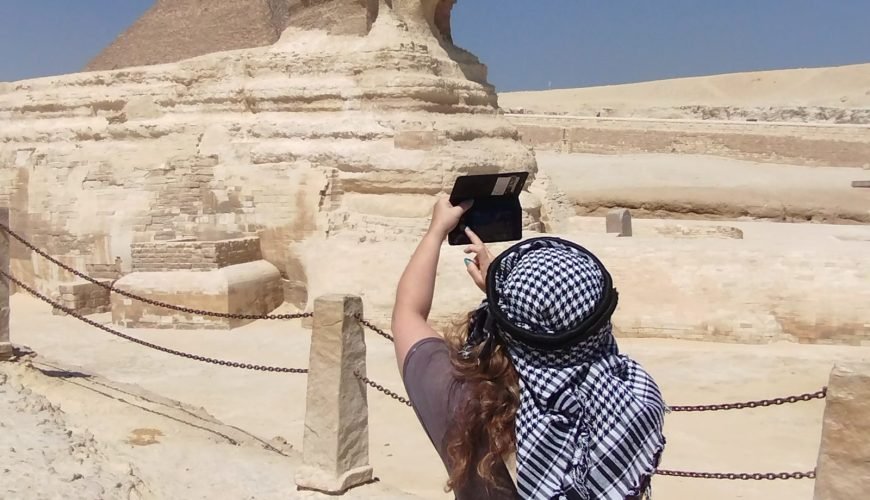 Une femme de dos portant un keffieh traditionnel photographie le Grand Sphinx de Gizeh avec son smartphone, une étape clé du voyage Pyramides depuis Sharm El-Sheikh