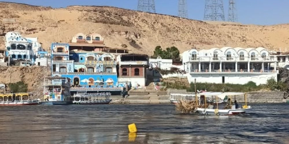 Vue sur les maisons colorées du village nubien sur les rives du Nil avec des felouques et des pylônes en arrière-plan, lors d'une Visite inoubliable a Souk du Assouan et village nubien