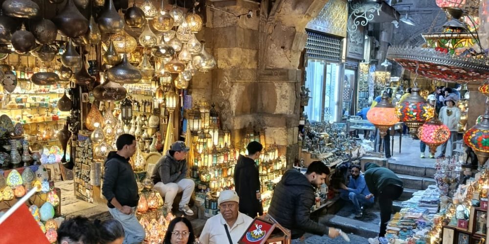 Une rue du souk Khan el-Khalili, en Voyage en Egypte, bordée de boutiques vendant des lanternes traditionnelles en verre coloré et des souvenirs artisanaux sous des arches en pierre historique.