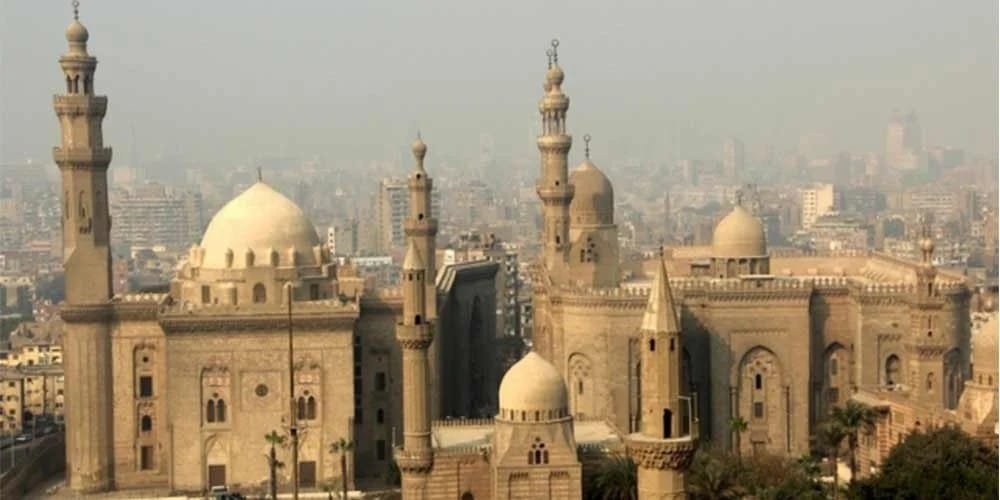 Vue panoramique sur la Mosquée-Madrasa du Sultan Hassan et la Mosquée Al-Rifa'i au Caire, avec leurs minarets imposants et leurs dômes sous un ciel légèrement brumeux, symbolisant un Voyage en Egypte historique.