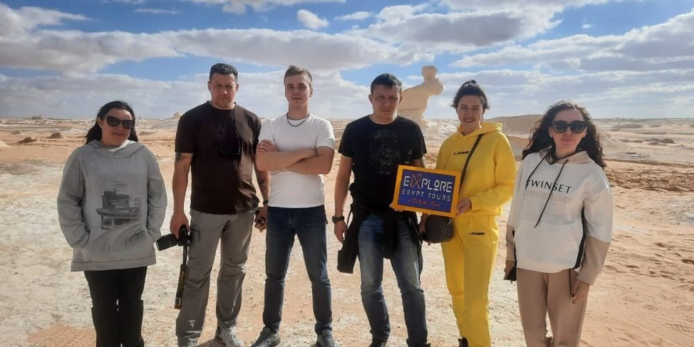 Grupo de seis turistas posando no Deserto Branco, com céu parcialmente nublado e formações rochosas ao fundo; uma mulher segura uma placa da Explore Egypt Tours.