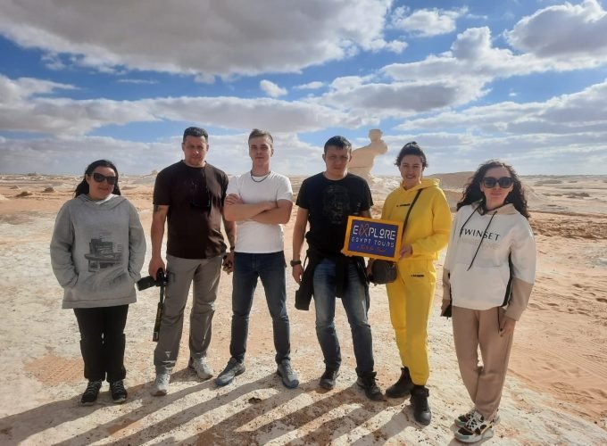 Grupo de seis turistas posando no Deserto Branco, com céu parcialmente nublado e formações rochosas ao fundo; uma mulher segura uma placa da Explore Egypt Tours.