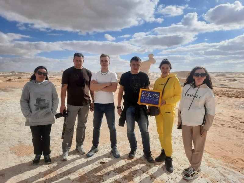 Grupo de seis turistas posando no Deserto Branco, com céu parcialmente nublado e formações rochosas ao fundo; uma mulher segura uma placa da Explore Egypt Tours.