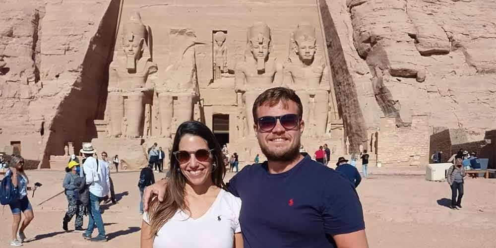 A medium shot of a smiling man and woman wearing sunglasses and casual t-shirts, standing in the foreground. Behind them is the massive rock-cut facade of the Great Temple of Ramesses II at Abu Simbel. The facade features four colossal seated statues of the Pharaoh, with the second statue from the left partially destroyed. Other tourists are visible in the wide, sunny courtyard under a clear blue sky. This iconic location is the highlight of an Abu Simbel sharing day tour