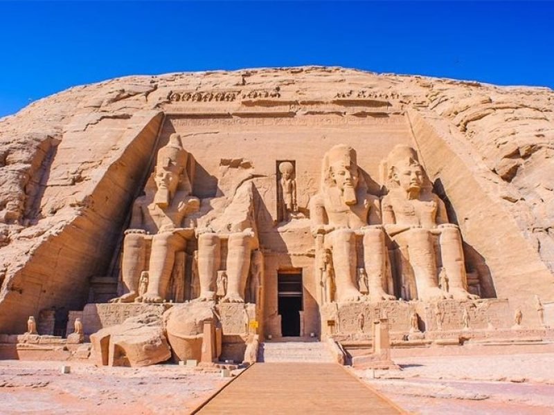 A full, direct wide shot of the Great Temple of Ramesses II at Abu Simbel carved into a massive sandstone cliff. The facade features four colossal seated statues of the Pharaoh, though the second statue from the left is partially collapsed with its head and torso lying on the ground in front. A central entrance leads into the temple, flanked by smaller standing statues of royal family members at the feet of the colossi. A wooden walkway in the foreground leads directly to the temple entrance under a vivid, cloudless blue sky. This iconic site is the main highlight of an Abu Simbel sharing day tour