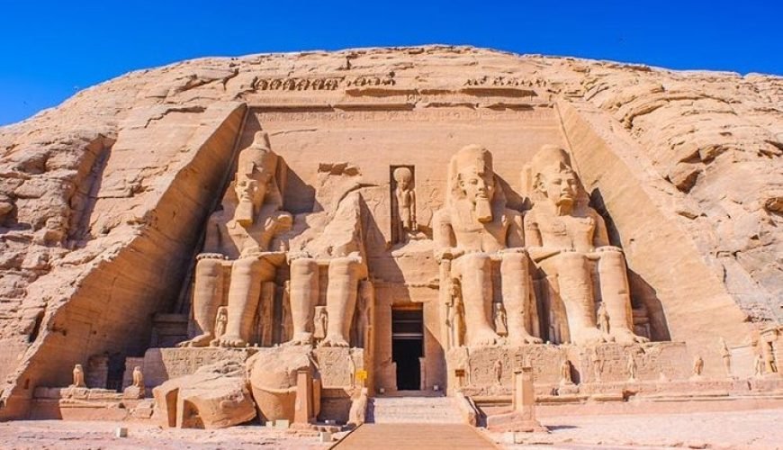 A full, direct wide shot of the Great Temple of Ramesses II at Abu Simbel carved into a massive sandstone cliff. The facade features four colossal seated statues of the Pharaoh, though the second statue from the left is partially collapsed with its head and torso lying on the ground in front. A central entrance leads into the temple, flanked by smaller standing statues of royal family members at the feet of the colossi. A wooden walkway in the foreground leads directly to the temple entrance under a vivid, cloudless blue sky. This iconic site is the main highlight of an Abu Simbel sharing day tour