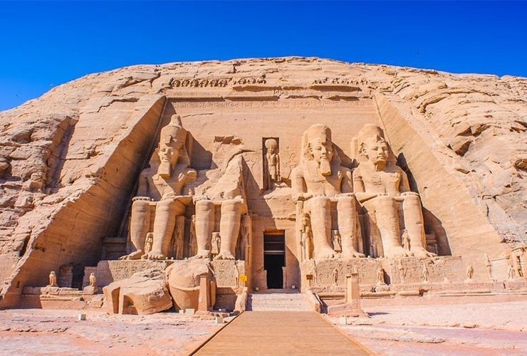 A full, direct wide shot of the Great Temple of Ramesses II at Abu Simbel carved into a massive sandstone cliff. The facade features four colossal seated statues of the Pharaoh, though the second statue from the left is partially collapsed with its head and torso lying on the ground in front. A central entrance leads into the temple, flanked by smaller standing statues of royal family members at the feet of the colossi. A wooden walkway in the foreground leads directly to the temple entrance under a vivid, cloudless blue sky. This iconic site is the main highlight of an Abu Simbel sharing day tour