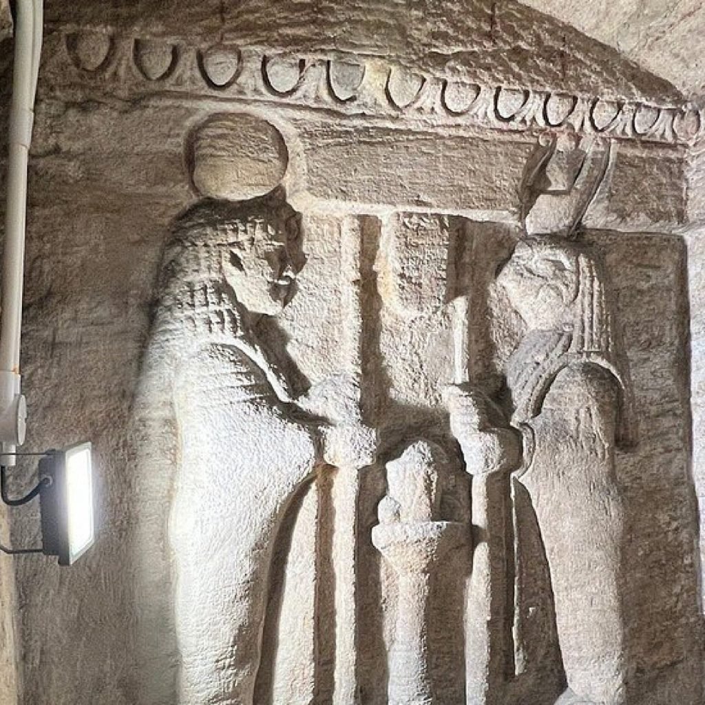 An ancient limestone wall carving inside the Catacombs of Kom El Shoqafa featuring two Pharaonic figures in ritual poses. The intricate relief shows traditional Egyptian headdresses and scepters illuminated by an artificial white light during an Alexandria Discovering from Alexandria Port excursion. The stone surface has a rough texture and historical patterns carved into the upper border of the burial chamber
