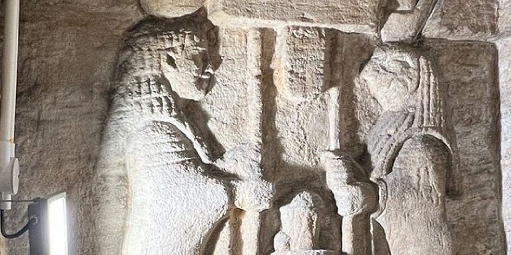 An ancient limestone wall carving inside the Catacombs of Kom El Shoqafa featuring two Pharaonic figures in ritual poses. The intricate relief shows traditional Egyptian headdresses and scepters illuminated by an artificial white light during an Alexandria Discovering from Alexandria Port excursion. The stone surface has a rough texture and historical patterns carved into the upper border of the burial chamber