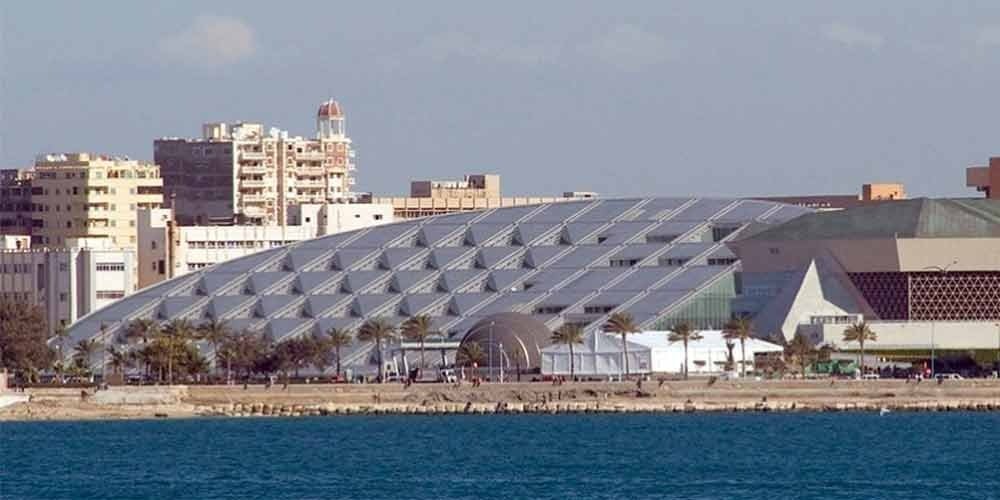 A panoramic view of the modern Bibliotheca Alexandrina featuring its iconic circular tilted glass roof near the Mediterranean coast. Lush palm trees line the waterfront walkway in front of the innovative architectural landmark during an Alexandria Discovering from Alexandria Port. The cityscape in the background shows various multi-story buildings under a clear, bright sky