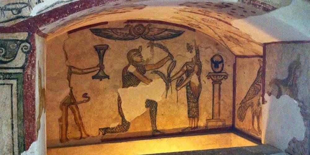 Une peinture murale antique à l'intérieur d'une chambre funéraire voûtée. On y voit des personnages de profil, dont un pharaon offrant des plumes à une divinité, sous un disque solaire ailé. Les couleurs sont dans les tons ocre, rouge et noir sur un fond beige lors d'une excursion Alexendrie dans une journée au départ du Caire