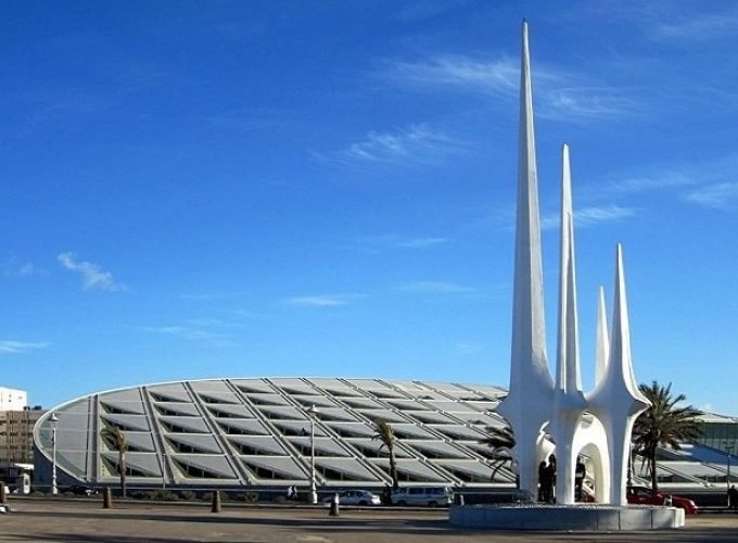Une vue extérieure de la bibliothèque d'Alexandrie avec son toit incliné en forme de disque argenté et ses murs en granit gravés. Au premier plan, on voit un monument blanc moderne composé de trois flèches hautes et pointues s'élevant vers un ciel bleu clair lors d'une excursion Alexendrie dans une journée au départ du Caire