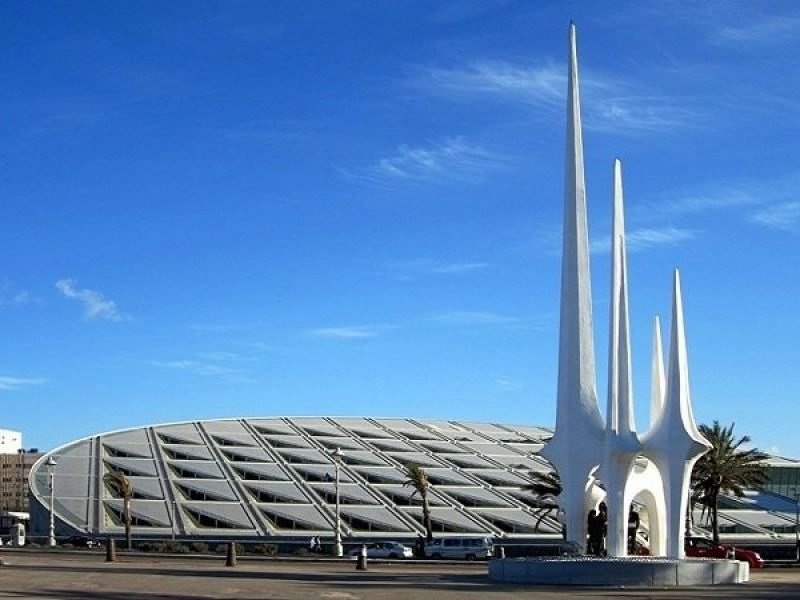 Une vue extérieure de la bibliothèque d'Alexandrie avec son toit incliné en forme de disque argenté et ses murs en granit gravés. Au premier plan, on voit un monument blanc moderne composé de trois flèches hautes et pointues s'élevant vers un ciel bleu clair lors d'une excursion Alexendrie dans une journée au départ du Caire
