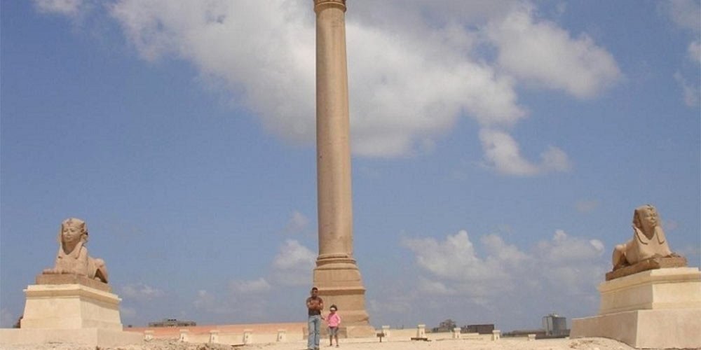 A wide shot of the towering Pompey's Pillar in Alexandria, flanked by two ancient sphinx statues on white pedestals under a blue sky with white clouds during an Egypt Vacation