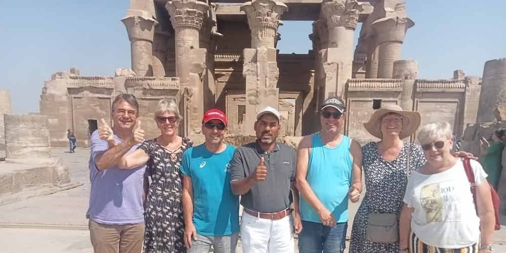 A group of six happy tourists and their local guide posing with thumbs up in front of the massive ancient stone columns of Kom Ombo Temple during an Aswan tour