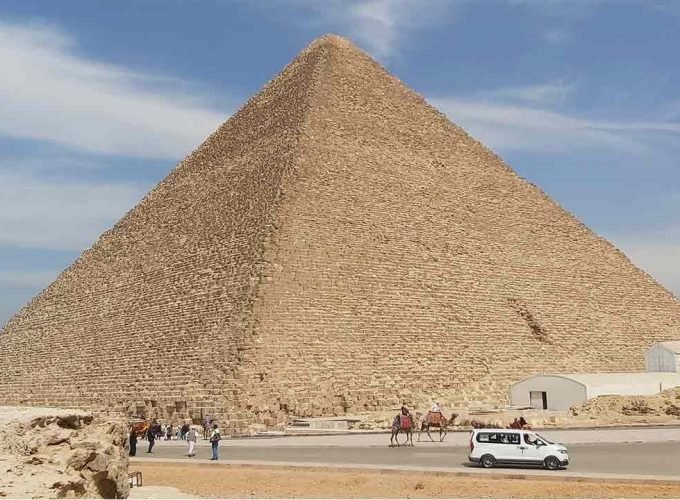 A massive ancient pyramid stands tall under a blue sky with light wispy clouds. At its base, tourists, camels with riders, and a white van move along a paved road during a Cairo Day Tour By Flight from Safaga & excursion