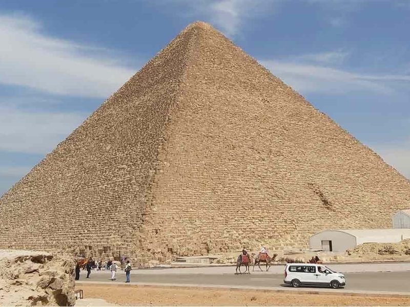 A massive ancient pyramid stands tall under a blue sky with light wispy clouds. At its base, tourists, camels with riders, and a white van move along a paved road during a Cairo Day Tour By Flight from Safaga & excursion