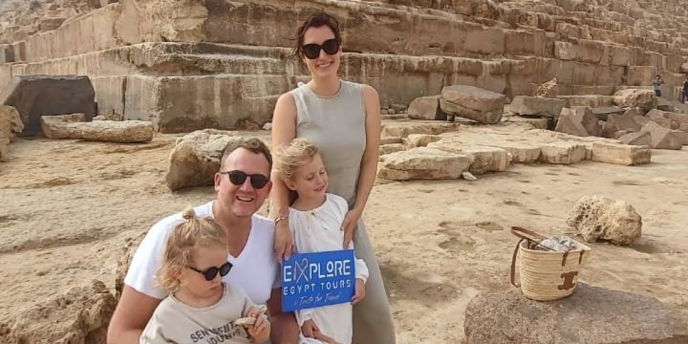 A happy family of four posing together in front of the massive stone blocks of a Great Pyramid at Giza. They are holding a small blue sign while sitting on ancient rocks during their Cairo Day Tour By Flight from Safaga
