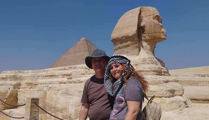 A medium-wide shot of a man and a woman posing together at the Giza Plateau. The man on the left wears a brown t-shirt and a grey sun hat, while the woman on the right wears a grey t-shirt, a beige backpack, and a black and white keffiyeh scarf on her head. Directly behind them is the Great Sphinx of Giza, shown in profile facing right. In the far background, the Great Pyramid of Giza rises into a clear, bright blue sky. A low stone wall with a dark metal chain fence is visible in the lower-left foreground, capturing a classic moment from a Cairo Day Trip for Groups