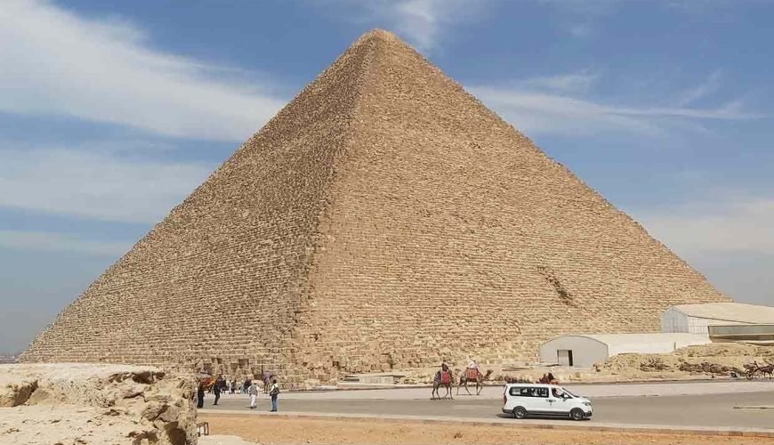A massive, detailed view of the Great Pyramid of Giza under a blue sky with light clouds. In the foreground, a paved road shows a white van driving by, people walking, and two tourists on camels. A small white modern building is nestled near the base of the ancient limestone structure.
