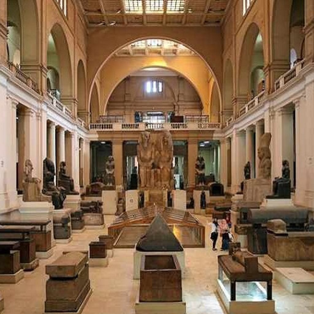 Interior view of the Egyptian Museum in Cairo showing a grand neoclassical hall with high arches, columns, statues of pharaohs, stone sarcophagi, ancient artifacts, display cases, and visitors walking through the central gallery – Cairo Adventures for Group
