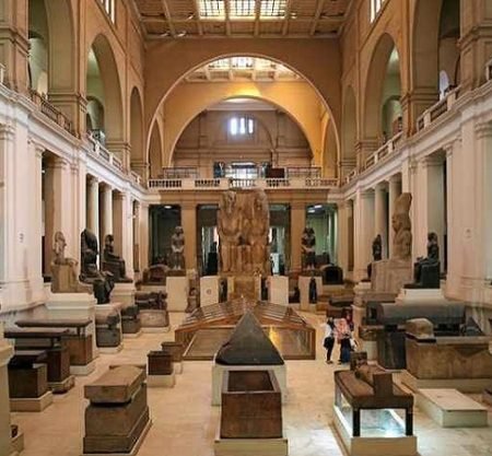 Interior view of the Egyptian Museum in Cairo showing a grand neoclassical hall with high arches, columns, statues of pharaohs, stone sarcophagi, ancient artifacts, display cases, and visitors walking through the central gallery – Cairo Adventures for Group