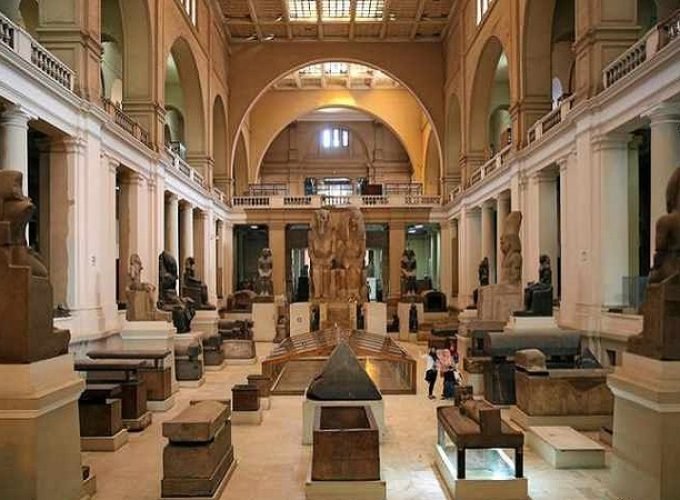 Interior view of the Egyptian Museum in Cairo showing a grand neoclassical hall with high arches, columns, statues of pharaohs, stone sarcophagi, ancient artifacts, display cases, and visitors walking through the central gallery – Cairo Adventures for Group