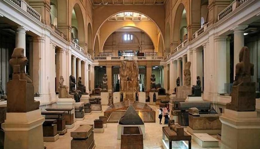 Interior view of the Egyptian Museum in Cairo showing a grand neoclassical hall with high arches, columns, statues of pharaohs, stone sarcophagi, ancient artifacts, display cases, and visitors walking through the central gallery – Cairo Adventures for Group