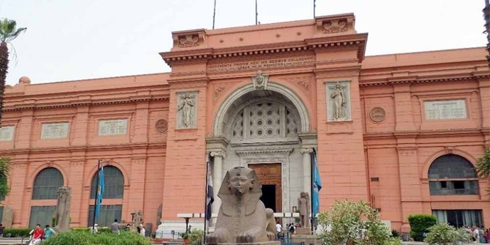 Exterior view of the Egyptian Museum in Cairo showing the pink neoclassical façade, the main arched entrance, carved stone reliefs, national flags, palm trees, visitors in front of the building, and a large sphinx statue placed at the entrance – Cairo Adventures for Grou