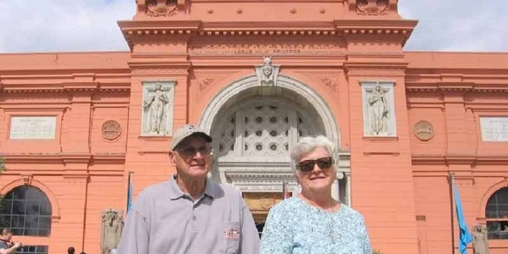 An elderly couple smiling in front of the pink Egyptian Museum in Cairo with three flags flying on top under a blue sky. The man wears a grey polo and cap, and the woman wears a light blue patterned shirt on a Tour from Marina Sharm