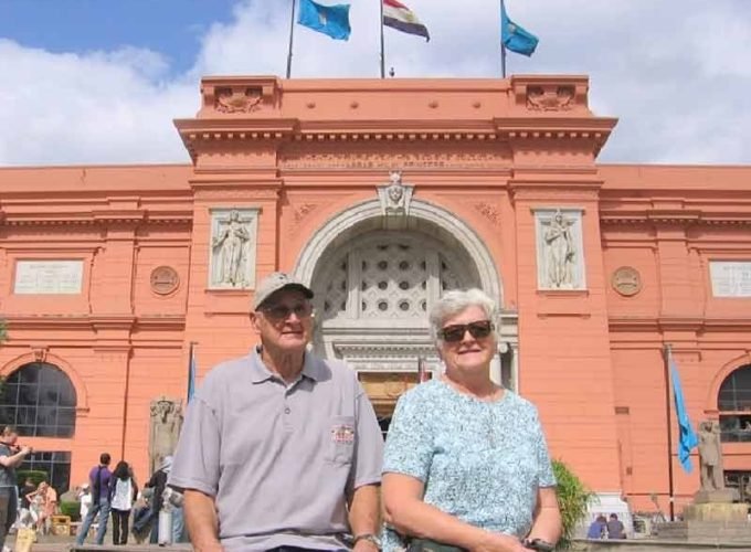 An elderly couple smiling in front of the pink Egyptian Museum in Cairo with three flags flying on top under a blue sky. The man wears a grey polo and cap, and the woman wears a light blue patterned shirt on a Tour from Marina Sharm