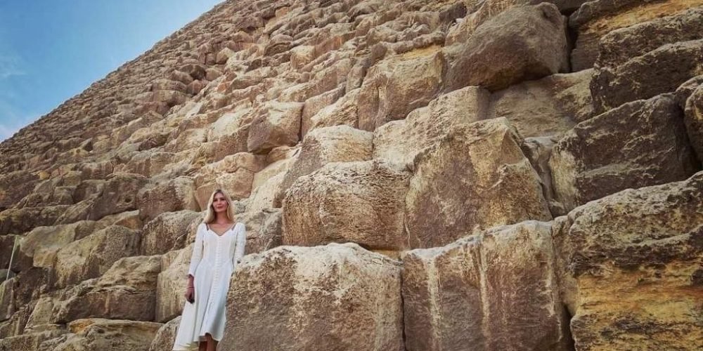 A low-angle shot of a woman in a white dress standing on the massive limestone blocks of the Great Pyramid's base. The ancient stone structure towers upward toward a bright blue sky with wispy clouds during an excursion from Sokhna Port