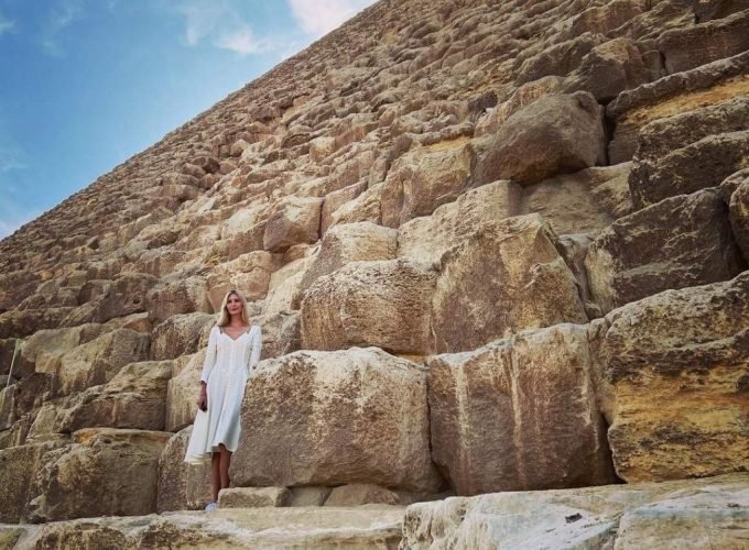 A low-angle shot of a woman in a white dress standing on the massive limestone blocks of the Great Pyramid's base. The ancient stone structure towers upward toward a bright blue sky with wispy clouds during an excursion from Sokhna Port