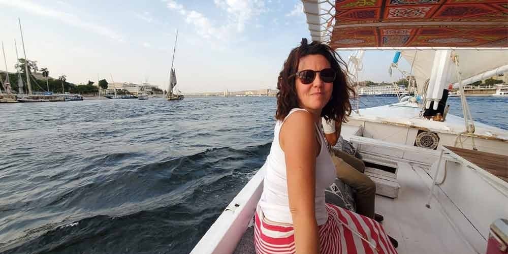 A woman in a white tank top and red striped skirt sitting on a traditional felucca boat sailing on the Nile River. The blue water is filled with other sailboats and city buildings in the distance under a bright sky during an excursion from Sokhna Port