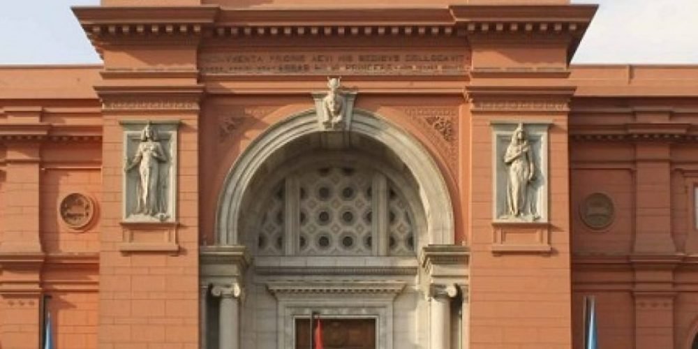 The grand red facade of the Egyptian Museum in Tahrir Square featuring a large arched entrance and classical statues on either side. The building is topped with three flags including the Egyptian national flag under a clear sky during a Cairo Trip. Detailed architectural carvings and stone columns frame the main doorway where tourists enter the historic landmark