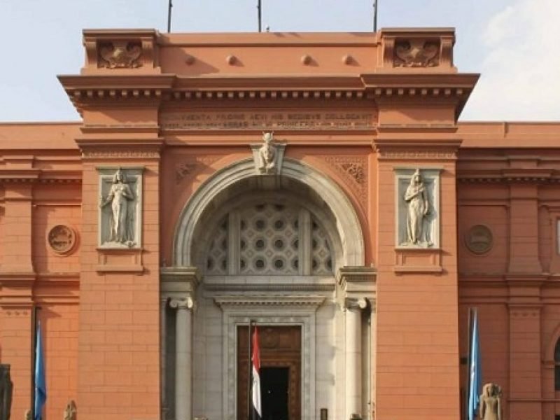 The grand red facade of the Egyptian Museum in Tahrir Square featuring a large arched entrance and classical statues on either side. The building is topped with three flags including the Egyptian national flag under a clear sky during a Cairo Trip. Detailed architectural carvings and stone columns frame the main doorway where tourists enter the historic landmark