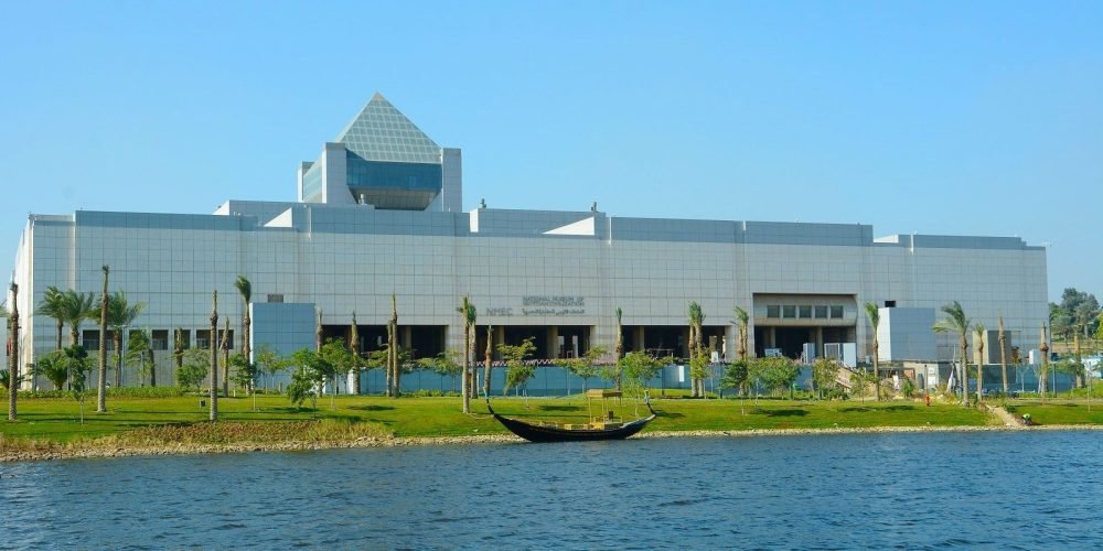 The National Museum of Egyptian Civilization beside a calm lake, surrounded by trees and green landscape