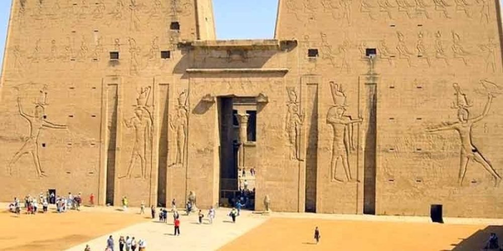 Une vue frontale d'un pylône égyptien massif en pierre beige avec une entrée centrale haute. Les murs sont décorés de gravures géantes représentant des divinités égyptiennes tenant des sceptres. Plusieurs touristes marchent sur l'esplanade sablonneuse devant le monument lors d'un Circuit au Caire Assouan Louxor et Hurghada