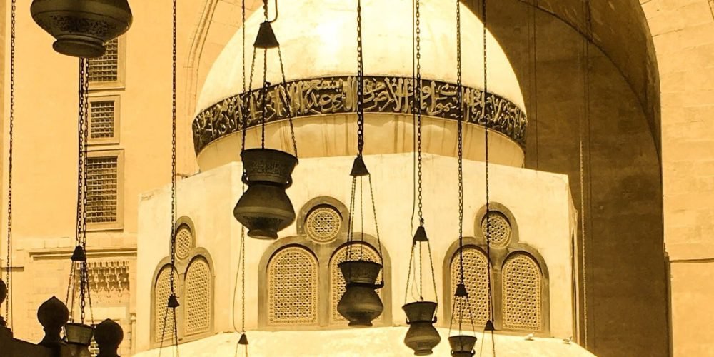 Une vue rapprochée d'une fontaine de purification avec un dôme blanc orné d'une calligraphie arabe foncée. Au premier plan, plusieurs lanternes en métal noir sont suspendues par de longues chaînes, devant une haute arche en pierre lors d'une visite au Caire Islamique