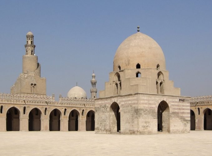 Une très large cour intérieure pavée de dalles claires sous un ciel bleu. Au centre se trouve un grand édifice carré en pierre avec des arches et un dôme imposant. Sur le côté, une muraille à arcades entoure la cour et un minaret en spirale unique s'élève en arrière-plan au Caire Islamique