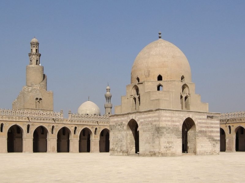 Une très large cour intérieure pavée de dalles claires sous un ciel bleu. Au centre se trouve un grand édifice carré en pierre avec des arches et un dôme imposant. Sur le côté, une muraille à arcades entoure la cour et un minaret en spirale unique s'élève en arrière-plan au Caire Islamique