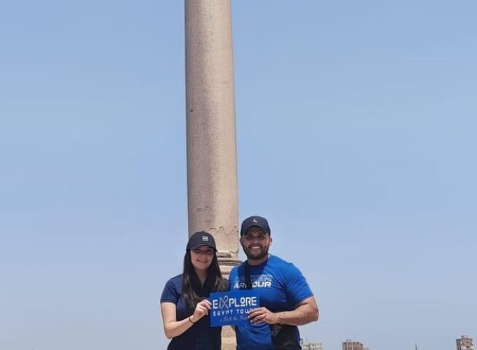 Un jeune homme et une jeune femme souriants، portant des casquettes noires et des t-shirts bleus، posent ensemble en tenant une petite pancarte bleue. Derrière eux se dresse l'immense colonne de Pompée en granit rouge qui s'élève vers un ciel bleu pur et sans nuages lors d'un circuit le Caire et Alexandrie