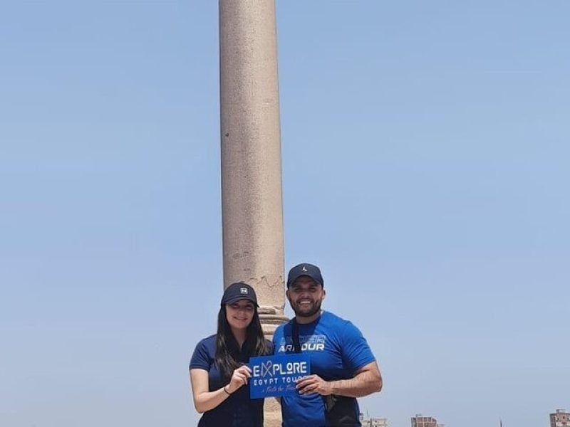 Un jeune homme et une jeune femme souriants، portant des casquettes noires et des t-shirts bleus، posent ensemble en tenant une petite pancarte bleue. Derrière eux se dresse l'immense colonne de Pompée en granit rouge qui s'élève vers un ciel bleu pur et sans nuages lors d'un circuit le Caire et Alexandrie