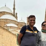 Un homme et une femme souriants posent ensemble devant un grand mur en pierre calcaire. En arrière-plan, on voit les dômes majestueux et les hauts minarets de la mosquée de Méhémet Ali sous un ciel couvert lors d'un circuit le Caire et Alexandrie