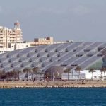 Une vue panoramique depuis la mer Méditerranée montrant le bâtiment moderne de la bibliothèque d'Alexandrie avec son grand toit circulaire incliné en métal gris. On voit des palmiers le long de la côte، des bâtiments urbains en arrière-plan et des eaux bleues au premier plan lors d'un circuit le Caire et Alexandrie