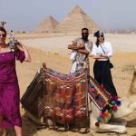 Trois touristes, deux hommes et une femme portant des lunettes de soleil et des chapeaux, sourient en se tenant debout dans le désert. À côté d'eux, un chameau est assis sur le sable avec une selle colorée. En arrière-plan, on voit les grandes pyramides de Gizeh sous un ciel bleu clair lors d'un voyage le Caire et Alexandrie