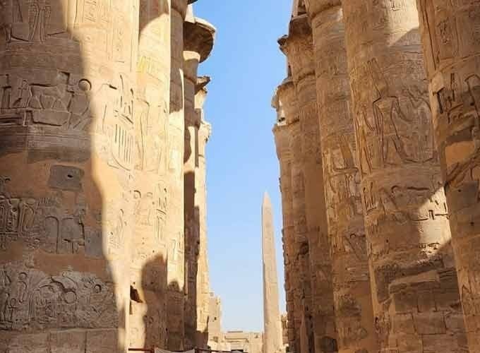 Une perspective entre deux rangées de colonnes de pierre massives et sculptées de hiéroglyphes égyptiens anciens. Au centre de l'allée، on aperçoit un grand obélisque pointu qui s'élève vers un ciel bleu clair sans nuages dans la ville de Louxor