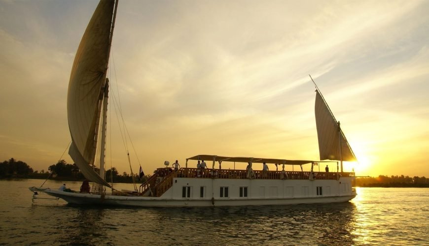 Une grande embarcation blanche de type Dahabiya avec deux voiles triangulaires déployées navigue sur le Nil. Le soleil se couche à l'horizon, créant une lumière dorée intense qui illumine l'eau et le ciel. On aperçoit des silhouettes de passagers sur le pont supérieur en bois